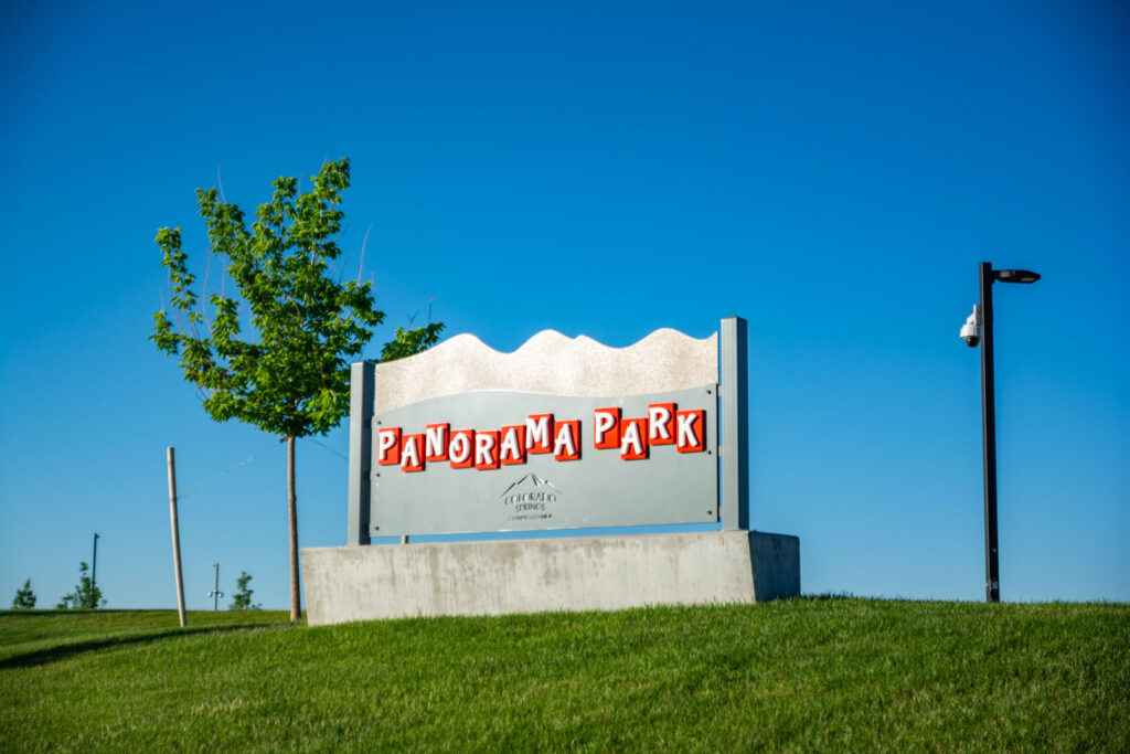 Photo of panorama park sign with blue skies and green grass. One tree and a camera